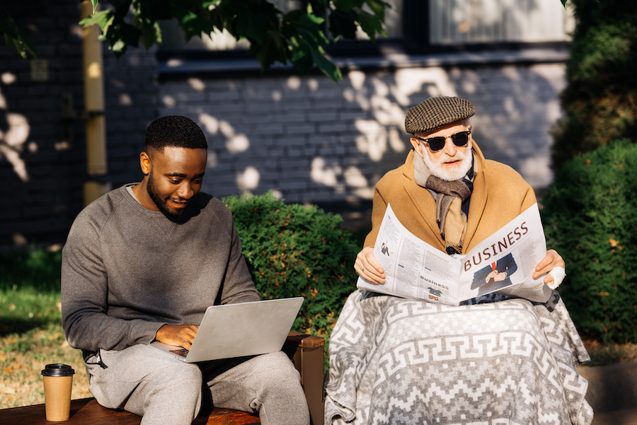 senior disabled man reading business newspaper in wheelchair while african american man using laptop