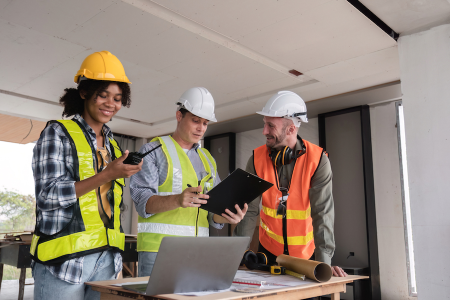 Engineering team in safety gear discusses work plan at a construction site, using a laptop and clipboard for planning and coordination.