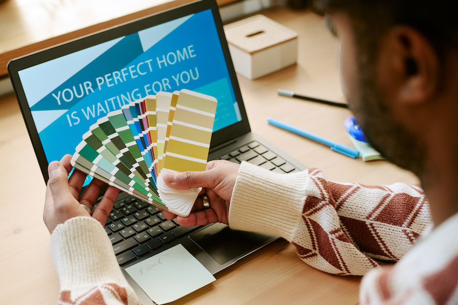 Young adult Black man holding color swatches while sitting at desk, selecting paint samples in front of laptop displaying home design message, working on interior project